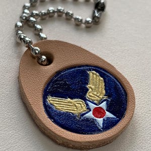 Puede incluir: Un llavero de cuero marrón con una cadena plateada. El llavero presenta un círculo azul con una estrella blanca y un centro rojo, rodeado de dos alas doradas.