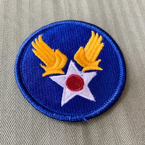 May include: A round, blue patch with a white star and a red center. Two yellow wings are above the star. The patch is embroidered with a blue border.