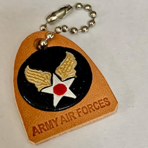 Op de afbeelding: Een bruinleren sleutelhanger met een zilveren kogelketting. De sleutelhanger heeft een zwarte cirkel met een witte ster en rood middelpunt, geflankeerd door gouden vleugels. De tekst "ARMY AIR FORCES" is in het leer geëmbosseerd.