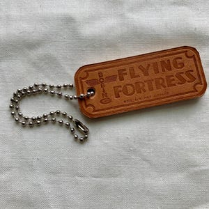 Könnte beinhalten: Ein rechteckiger, brauner Leder-Schlüsselanhänger mit dem geprägten Schriftzug "FLYING FORTRESS". Der Schlüsselanhänger hat eine silberne Kugelkette. Der Text "BOEING" ist ebenfalls eingeprägt. Der Hintergrund ist ein weißes, strukturiertes Gewebe.