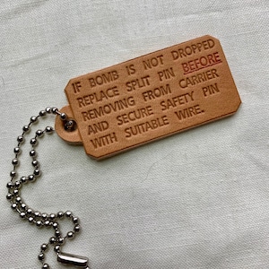 Könnte beinhalten: Ein rechteckiger, hellbrauner Lederanhänger mit geprägtem Text und einer silbernen Kugelkette. Der Text lautet: "IF BOMB IS NOT DROPPED REPLACE SPLIT PIN BEFORE REMOVING FROM CARRIER AND SECURE SAFETY PIN WITH SUITABLE WIRE."