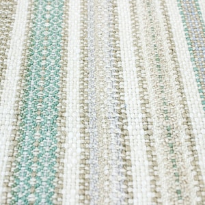 May include: Close-up of a woven rug with a striped pattern in shades of beige, white, and light green.