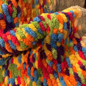 May include: A close-up of a colorful, textured blanket. The blanket features a vibrant mix of colors, including red, orange, yellow, green, blue, and purple. The material appears soft and plush, with a thick, looped texture. The blanket is folded over.