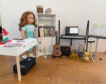 May include: A doll stands near a white desk with a red lamp and books. A miniature music studio includes a ukulele, keyboard, xylophone, and trumpet. A black case rests under the desk, and a white cabinet holds books.