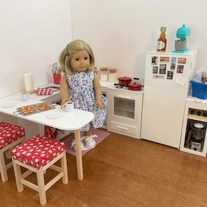 May include: A miniature kitchen scene featuring a doll in a blue floral dress. The set includes a white table, two stools with red and white patterned cushions, a stove, and a fridge with accessories.