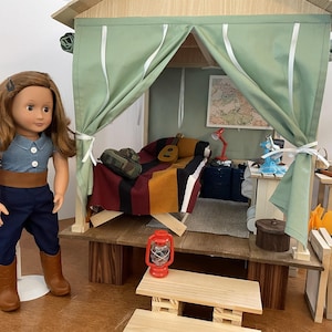 May include: A doll stands beside a miniature wooden playhouse with a green canvas roof and curtains. Inside, a bed with a colourful blanket, a guitar, and a map are visible. A red lantern and a small picnic table are in the foreground.