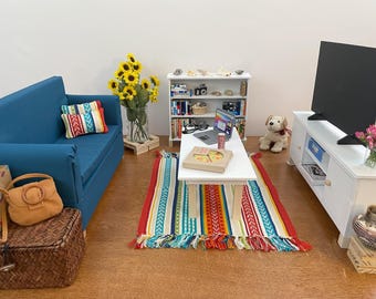 May include: A miniature living room scene with a blue sofa, colorful rug, and white coffee table. A TV, bookshelf, and decorative items complete the setting. A plush dog sits nearby.