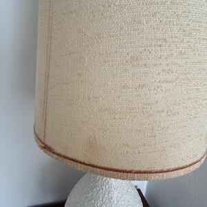 May include: A white ceramic table lamp with a textured, bumpy surface. The lamp has a beige fabric lampshade with a brown trim.