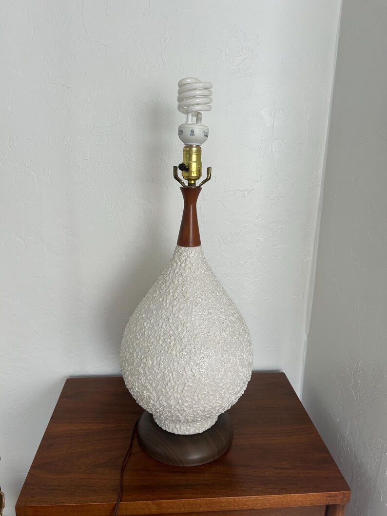May include: A white ceramic table lamp with a textured surface and a brown wooden base. The lamp has a white, spiral-shaped light bulb.