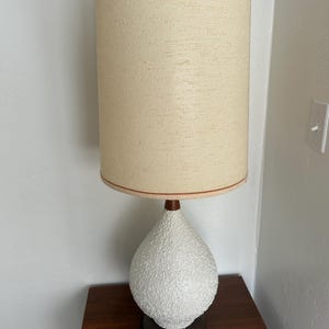 May include: A white ceramic table lamp with a textured finish and a brown wooden base. The lamp has a beige fabric shade with a brown trim.