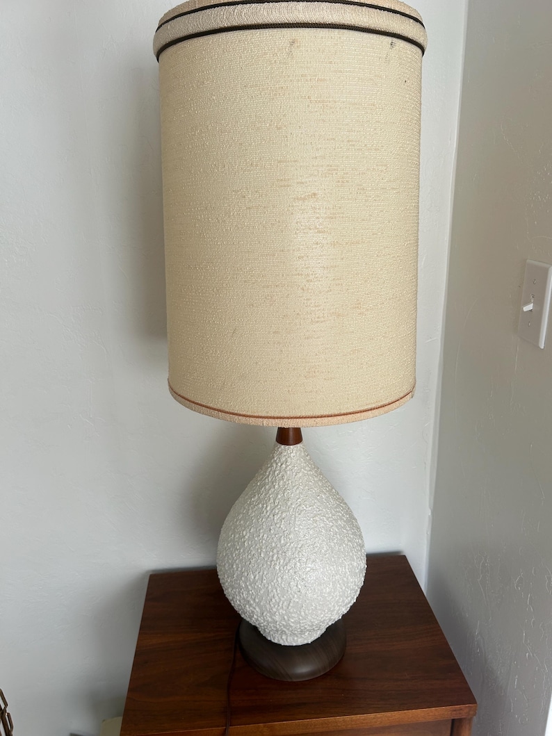 May include: A white ceramic table lamp with a textured finish and a brown wooden base. The lamp has a beige fabric shade with a brown trim.