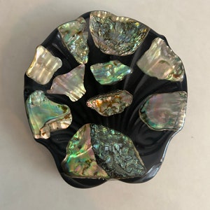May include: A black, scalloped dish with iridescent abalone shell inlay. The dish is perfect for holding jewelry, trinkets, or other small items.