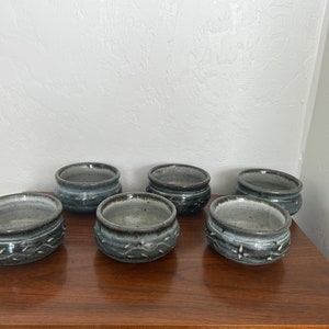 May include: Six small, grey and black ceramic bowls with a textured, speckled glaze. The bowls are arranged in two rows of three on a wooden surface.