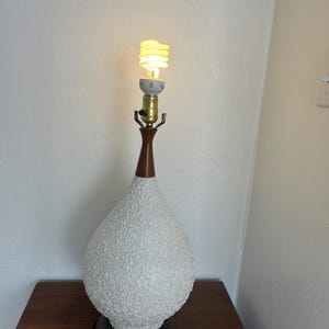May include: White ceramic table lamp with a textured surface and a wooden base. The lamp has a light bulb turned on.