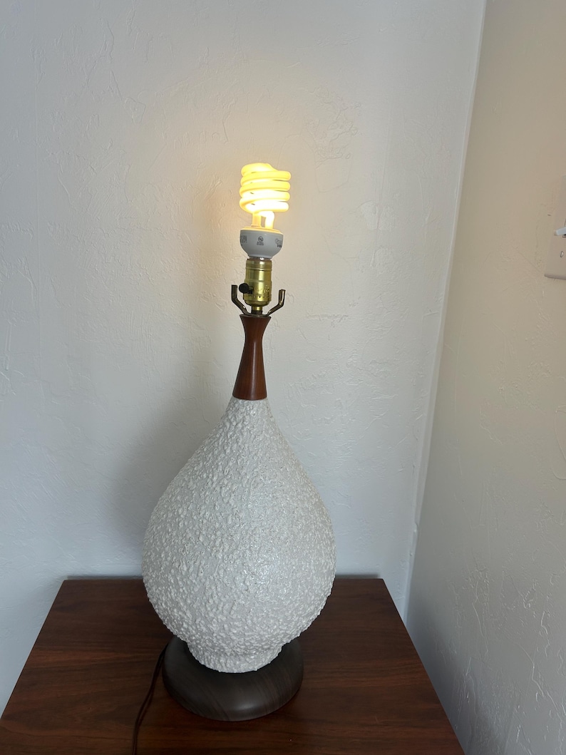 May include: White ceramic table lamp with a textured surface and a wooden base. The lamp has a light bulb turned on.