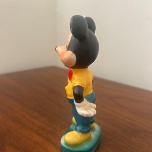 May include: A vintage Mickey Mouse figurine. The figure has a black head with large ears, a yellow shirt, blue pants, and yellow shoes. It stands on a green base and has white gloves and a red bow tie.