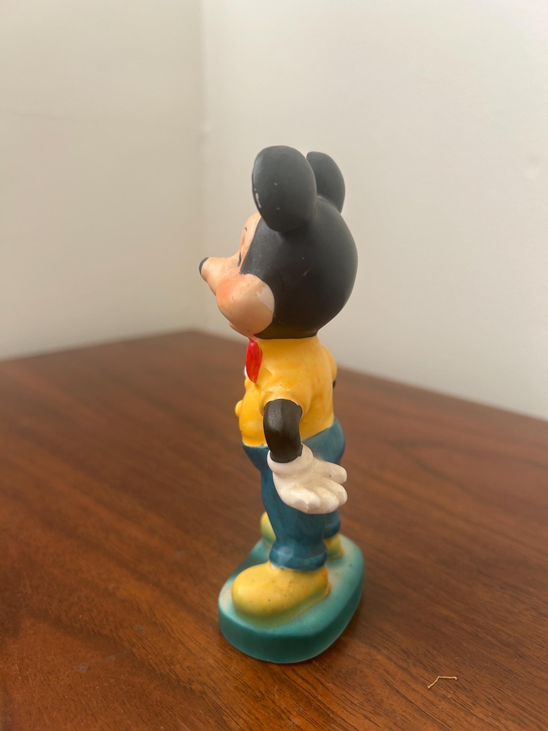 May include: A vintage Mickey Mouse figurine. The figure has a black head with large ears, a yellow shirt, blue pants, and yellow shoes. It stands on a green base and has white gloves and a red bow tie.