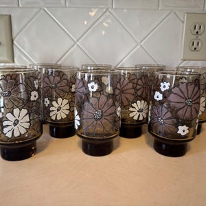May include: Set of eight vintage drinking glasses. Each glass features a floral design in shades of brown, white, and gray. The glasses have a dark brown base and a clear body. Perfect for serving water, juice, or cocktails.