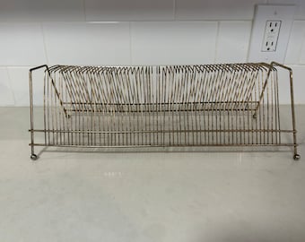 May include: A gold metal record rack with a grid design. The rack is empty and has a slight curve to it.