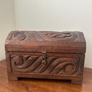May include: A small, antique wooden chest with a dark brown finish. The chest features intricate carved floral and swirl designs on the front, top, and sides. It has a hinged lid and a small metal clasp for closure. The chest is rectangular with a curved top.