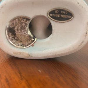 May include: A vintage ceramic figurine base with a light blue rim. The base has a hole and a silver-colored sticker with text. A small oval sticker reads "&copy; 1960 WALT DISNEY PRODUCTIONS".