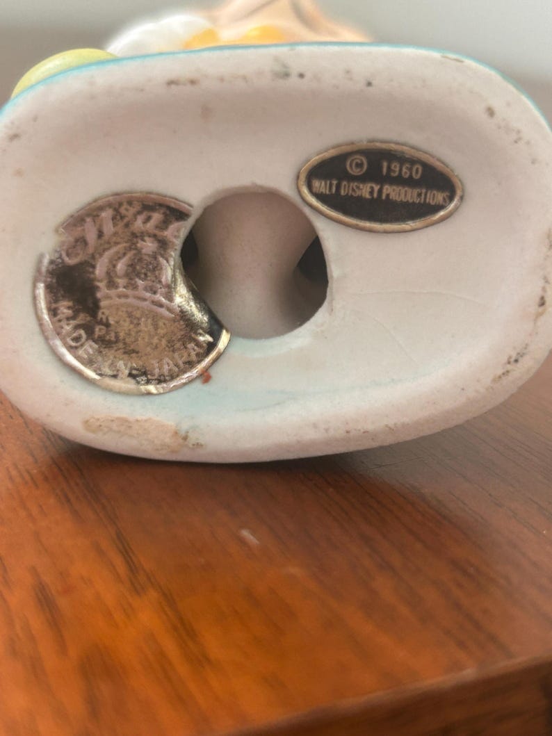 May include: A vintage ceramic figurine base with a light blue rim. The base has a hole and a silver-colored sticker with text. A small oval sticker reads "&copy; 1960 WALT DISNEY PRODUCTIONS".