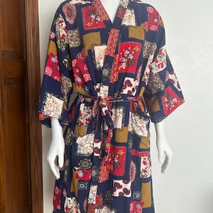 May include: A navy blue kimono-style robe with a patchwork design. The robe features a V-neck, wide sleeves, and a matching belt. The patchwork includes red, white, gold, and brown floral and geometric patterns. The robe is displayed on a mannequin.