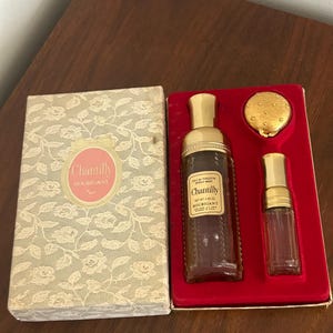 May include: A Houbigant Chantilly perfume gift set featuring a rectangular box with a cream lace pattern and a red velvet interior. The set includes a 2.50 oz spray mist bottle, a smaller glass vial, and a round gold compact, all with gold caps.