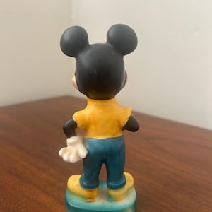 May include: A ceramic Mickey Mouse figurine, viewed from the back. The figure has a black head with large ears, a yellow shirt, blue pants, and yellow shoes. The figure is standing on a blue and yellow base.