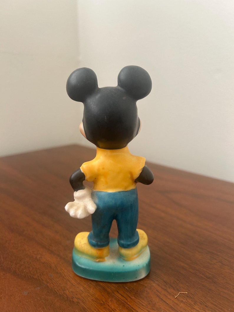 May include: A ceramic Mickey Mouse figurine, viewed from the back. The figure has a black head with large ears, a yellow shirt, blue pants, and yellow shoes. The figure is standing on a blue and yellow base.