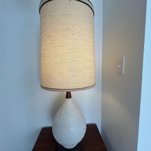 May include: A white ceramic table lamp with a textured finish and a brown wooden base. The lamp has a white fabric shade with a brown trim.