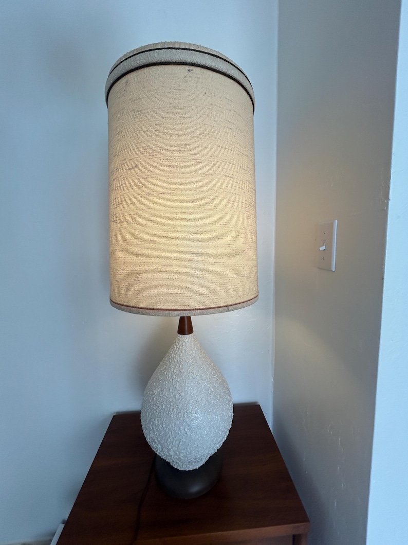 May include: A white ceramic table lamp with a textured finish and a brown wooden base. The lamp has a white fabric shade with a brown trim.