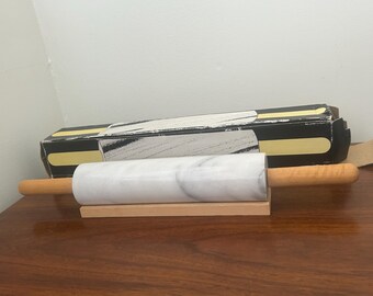 May include: A marble rolling pin with wooden handles rests on a wooden base. The rolling pin is white with gray veining. A black and gold box is in the background. Kitchen and baking supplies.