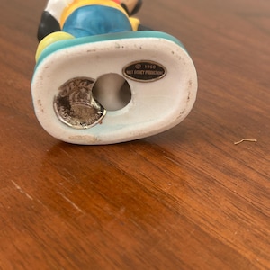 May include: A vintage Mickey Mouse figurine. The figurine is wearing a yellow shirt, blue shorts, and black gloves. The base is white and has a teal border. The bottom of the figurine has a stamp that reads "&copy; 1960 Walt Disney Productions" and "Made in England."