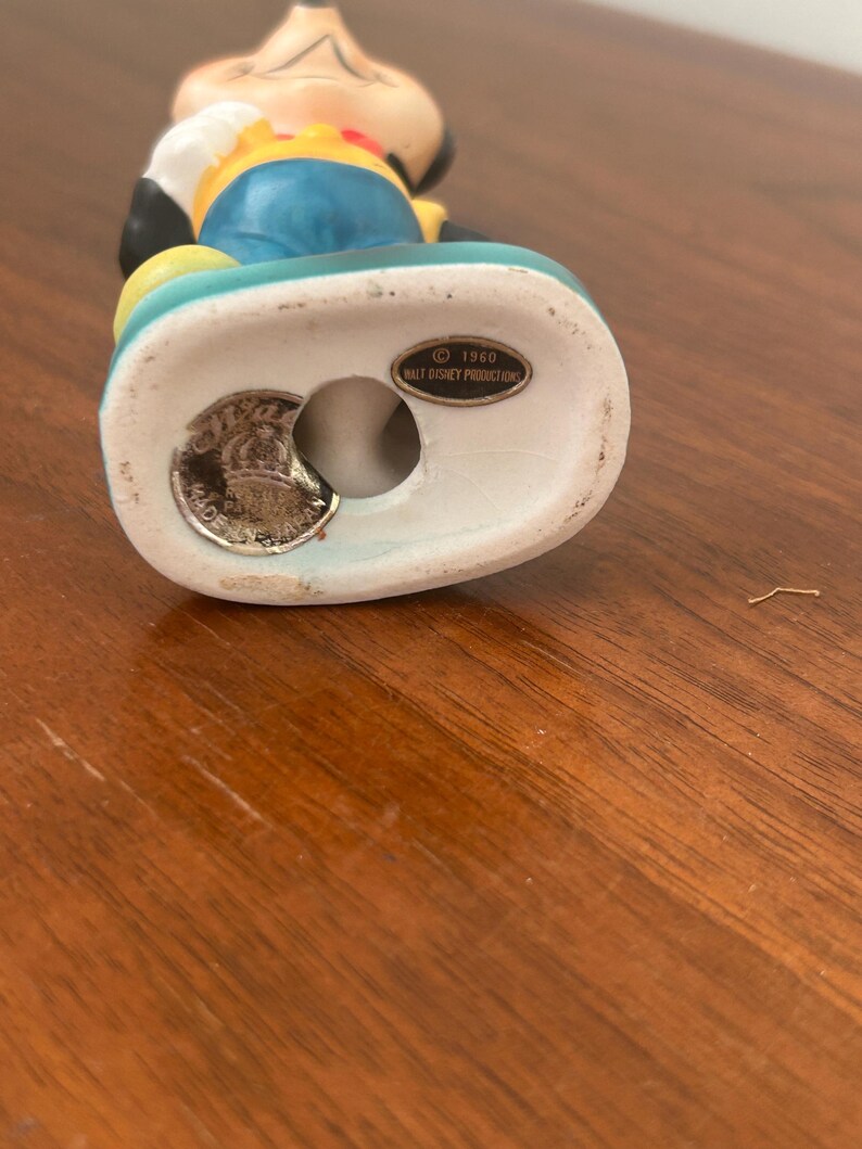 May include: A vintage Mickey Mouse figurine. The figurine is wearing a yellow shirt, blue shorts, and black gloves. The base is white and has a teal border. The bottom of the figurine has a stamp that reads "&copy; 1960 Walt Disney Productions" and "Made in England."