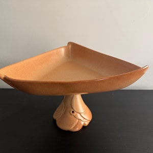 May include: A peach-colored, triangular serving dish with a pedestal base. The dish has a smooth, glossy finish and a slightly raised rim. The base is decorated with a floral design. The dish is likely made of ceramic or porcelain.