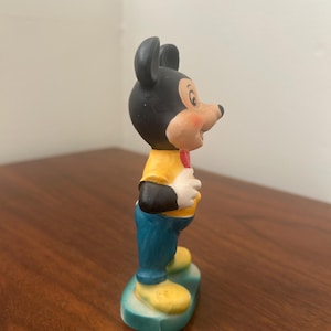 May include: A ceramic figurine of Mickey Mouse. The figure has a black head and ears, a yellow shirt, blue pants, and yellow shoes. A red bow tie is visible. The figurine is standing on a green base.