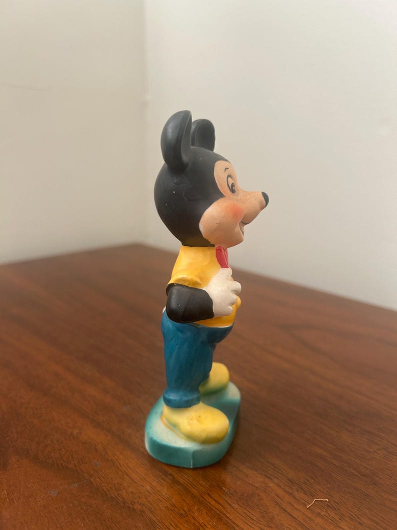 May include: A ceramic figurine of Mickey Mouse. The figure has a black head and ears, a yellow shirt, blue pants, and yellow shoes. A red bow tie is visible. The figurine is standing on a green base.
