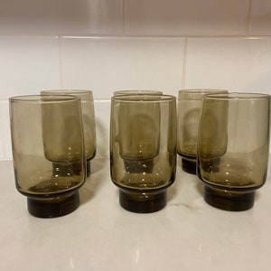 May include: Set of six vintage-style, brown-tinted drinking glasses. Each glass has a cylindrical shape with a slightly flared base. The glasses are arranged on a white countertop, showcasing their simple, yet elegant design.