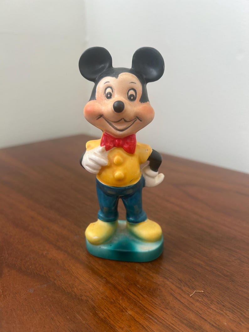 May include: A vintage Mickey Mouse figurine. The figure has black ears, a smiling face, and is wearing a yellow shirt, blue pants, a red bow tie, and yellow shoes. The figurine is standing on a green base.