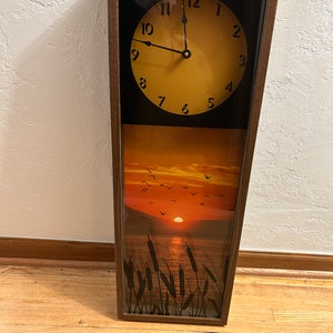 May include: A rectangular wall clock with a brown wooden frame. The clock face is yellow with black numbers and hands. The clock is set to 11:00. The background image is a sunset over a lake with birds flying in the sky.