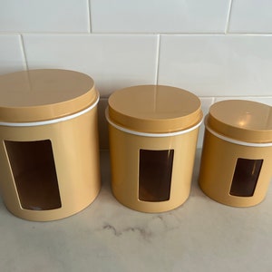 May include: Set of three yellow ceramic canisters with white lids and clear glass windows. The canisters are different sizes and are arranged in a row from largest to smallest.