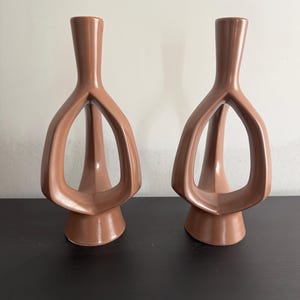 May include: Two brown ceramic vases with a unique, open design. The vases have a slender neck and a flared base, with an open, geometric shape in the middle. The vases are displayed on a dark surface against a white wall.