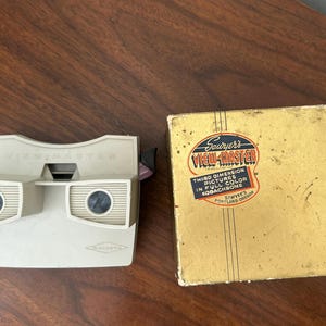 May include: A vintage View-Master viewer and its original box. The viewer is off-white with the word "VIEWMASTER" printed on the top. The box is gold with the words "VIEW-MASTER" and "THIRD DIMENSION PICTURES" printed on it.