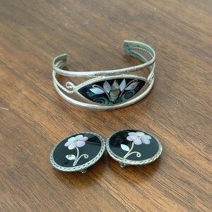 May include: A silver cuff bracelet and matching earrings set. The bracelet features an oval black inlay with iridescent shell accents. The earrings are round with black inlays and a white flower design.