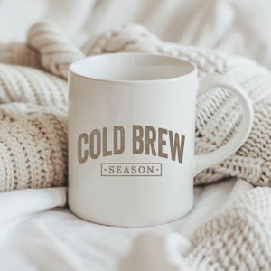 May include: A white ceramic mug featuring the text COLD BREW SEASON in a brown, distressed font. The mug has a smooth, glossy finish, a rounded handle on the right side, and a cylindrical body. The text is centered with the word SEASON inside a box.