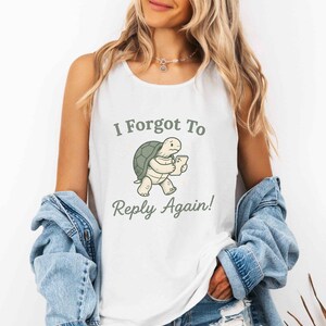 May include: A white sleeveless vest top featuring a graphic of a green turtle holding a smartphone. The text above the turtle reads I Forgot To and the text below reads Reply Again! in a dark green font. The garment is worn with a blue denim jacket.