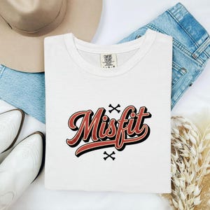 May include: A white short-sleeve t-shirt featuring the word Misfit in a red cursive font with a black outline. The design includes two small black crossbones symbols above and below the text. The shirt has a Comfort Colors brand label.