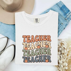 May include: A white short-sleeve t-shirt featuring the word TEACHER repeated four times in a stacked, wavy font. The text colors are orange, yellow, green, and blue, with small orange apple illustrations interspersed between the words.