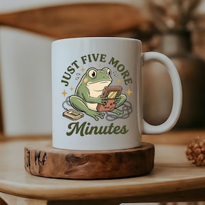 May include: A white ceramic mug featuring a green frog illustration holding a handheld gaming console with a cord. The text "JUST FIVE MORE Minutes" is printed in dark green arched lettering above and below the frog, with small yellow stars.